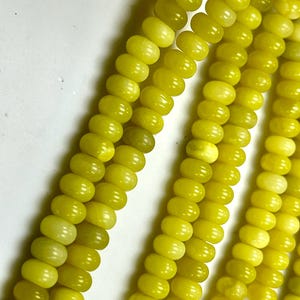 May include: Close-up of strands of yellow-green beads. The beads are oval-shaped and have a smooth, polished surface. The color is a vibrant, sunny yellow-green, with slight variations in tone. The beads are strung together, creating a flowing, textured appearance.