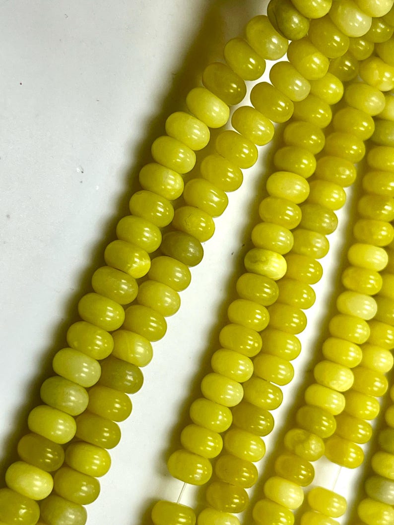 May include: Close-up of strands of yellow-green beads. The beads are oval-shaped and have a smooth, polished surface. The color is a vibrant, sunny yellow-green, with slight variations in tone. The beads are strung together, creating a flowing, textured appearance.