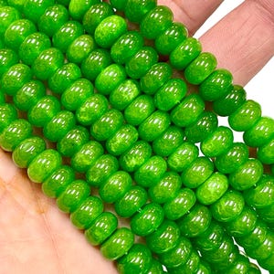 May include: Close-up of strands of bright green beads. The beads are round with a slightly flattened shape and a glossy finish. The beads are held in a hand, showcasing their vibrant colour and texture. Ideal for jewellery making or crafting.