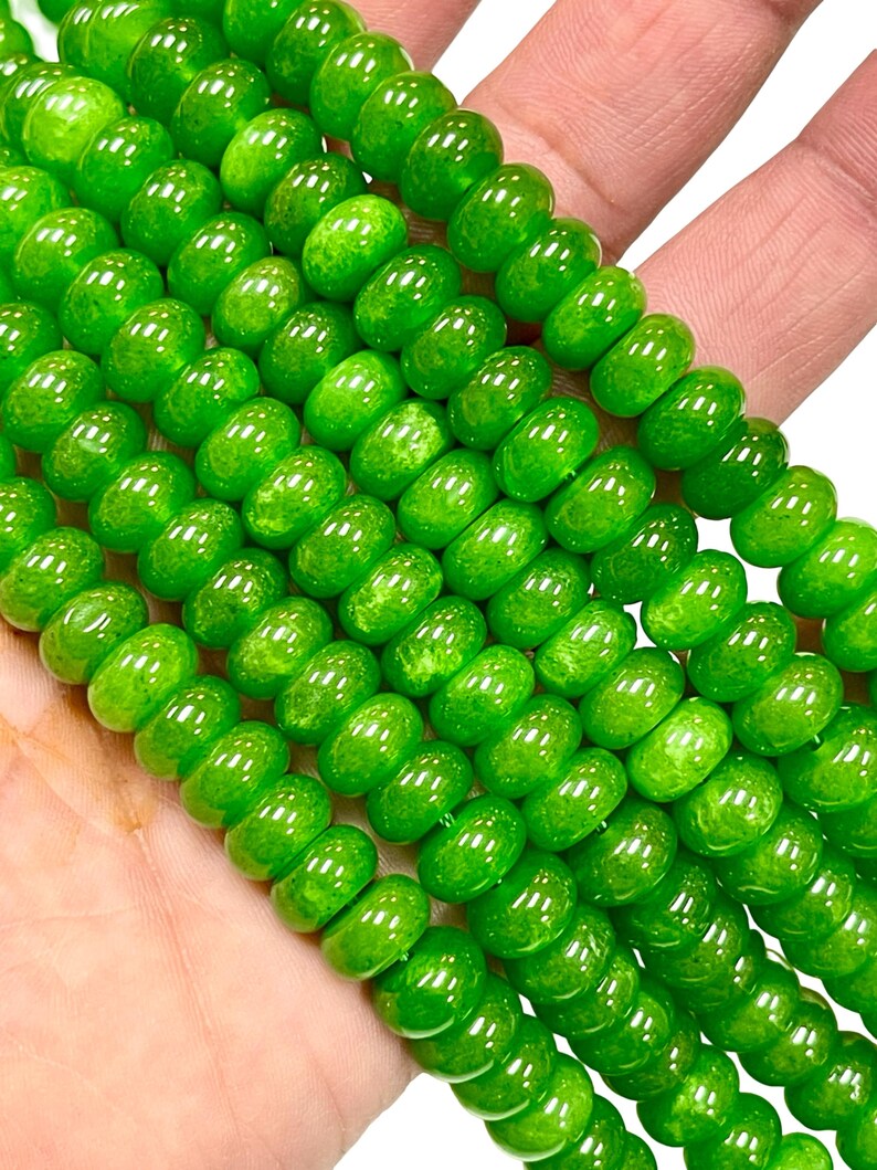 May include: Close-up of strands of bright green beads. The beads are round with a slightly flattened shape and a glossy finish. The beads are held in a hand, showcasing their vibrant colour and texture. Ideal for jewellery making or crafting.