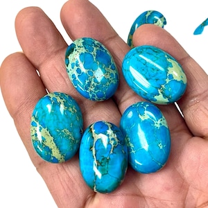 May include: A hand holding six oval-shaped gemstones. The stones have a vibrant blue base color with a mottled pattern of lighter blue and green. The gemstones are smooth and polished, reflecting light.