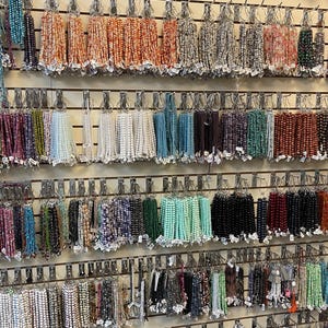 Puede incluir: Una pared de exhibici&oacute;n con varias hebras de cuentas de diferentes colores y materiales. Las cuentas est&aacute;n organizadas por color y tipo, y est&aacute;n colgadas de ganchos en una pared de madera.