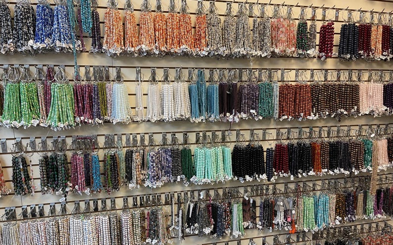 Puede incluir: Una pared de exhibici&oacute;n con varias hebras de cuentas de diferentes colores y materiales. Las cuentas est&aacute;n organizadas por color y tipo, y est&aacute;n colgadas de ganchos en una pared de madera.