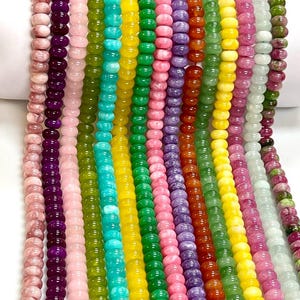 Op de afbeelding: Meerdere verticale strengen van gladde, ronde edelsteenkralen in een levendig kleurenspectrum, waaronder roze, paars, lichtroze, olijfgroen, turkoois, geel, felgroen, lichtroze, lavendel, oranje, groen, geel, magenta, wit en gevlekt groen.