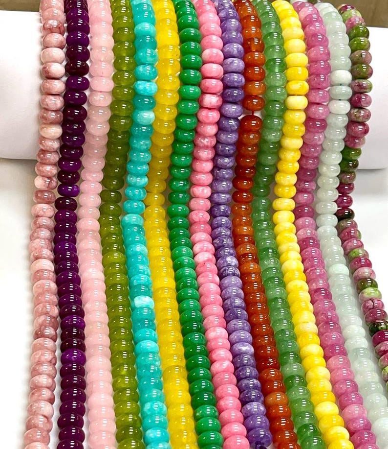 Op de afbeelding: Meerdere verticale strengen van gladde, ronde edelsteenkralen in een levendig kleurenspectrum, waaronder roze, paars, lichtroze, olijfgroen, turkoois, geel, felgroen, lichtroze, lavendel, oranje, groen, geel, magenta, wit en gevlekt groen.