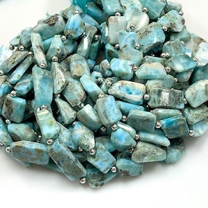 May include: A close-up of a necklace made of irregular, light blue and gray larimar stones. The stones are separated by small, silver-colored beads. The necklace is on a white background.