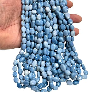 May include: A strand of oval-shaped, light blue beads. The beads have a smooth, polished surface and vary in shades of blue. The beads are strung together, creating a flowing, organic shape. The beads are held in a hand against a white background.