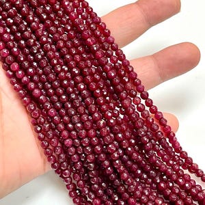 May include: Strands of faceted, deep red gemstone beads. The beads are small, round, and have a sparkling, polished finish. The beads are held in a hand, showcasing their color and texture. Ideal for jewelry making.
