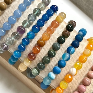 May include: A collection of multiple beaded bracelets arranged in rows, featuring smooth, polished spherical beads in a variety of colors including blue, orange, yellow, green, brown, and pink, displayed on a light-colored fabric stand.