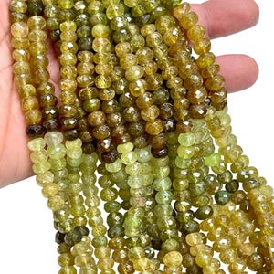 May include: Strands of faceted, olive-green gemstone beads. The beads vary in shade from light yellow-green to deep brown-green. The beads are strung together, creating a textured and colorful display. The beads are likely used for jewelry making.