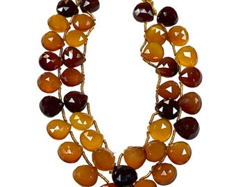May include: Two strands of faceted gemstone beads in shades of amber, brown, and yellow, with gold-colored stringing. The strands are topped with bright yellow tassels. The beads have a teardrop shape and are arranged in a symmetrical pattern.