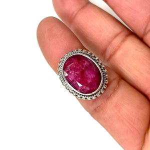 May include: A silver ring with a large oval-shaped ruby gemstone. The ruby is a deep red color and has a faceted cut. The ring is set in a silver band with a decorative border.
