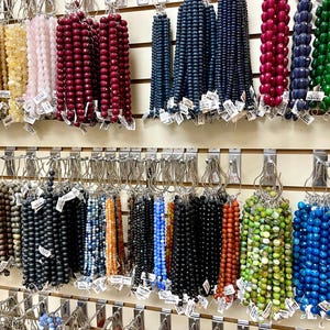 Puede incluir: Una pared con una exhibici&oacute;n de hilos de cuentas de varios colores, colgados de ganchos. Las cuentas est&aacute;n hechas de diferentes materiales, incluyendo vidrio, piedra y madera. Las cuentas est&aacute;n dispuestas en filas, con cada fila que contiene un color o tipo de cuenta diferente.