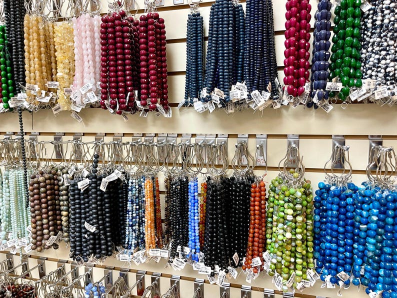 Puede incluir: Una pared con una exhibici&oacute;n de hilos de cuentas de varios colores, colgados de ganchos. Las cuentas est&aacute;n hechas de diferentes materiales, incluyendo vidrio, piedra y madera. Las cuentas est&aacute;n dispuestas en filas, con cada fila que contiene un color o tipo de cuenta diferente.