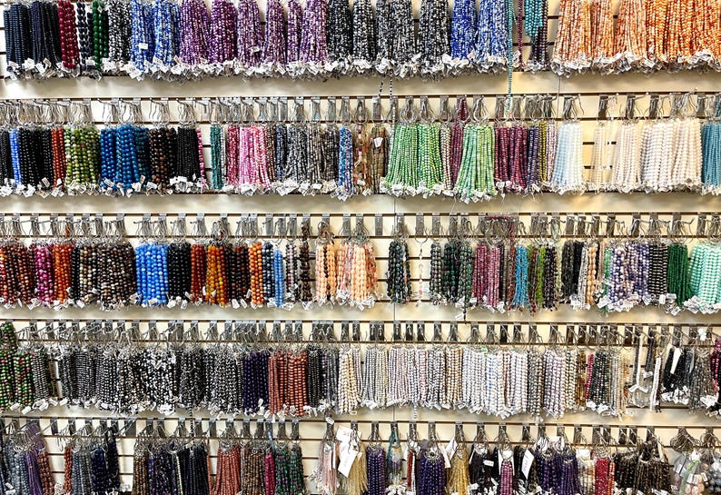 Puede incluir: Una pared de exhibici&oacute;n de varias hebras de cuentas de colores, organizadas en filas sobre un fondo blanco. Las cuentas est&aacute;n hechas de diferentes materiales, incluyendo vidrio, piedra y metal.