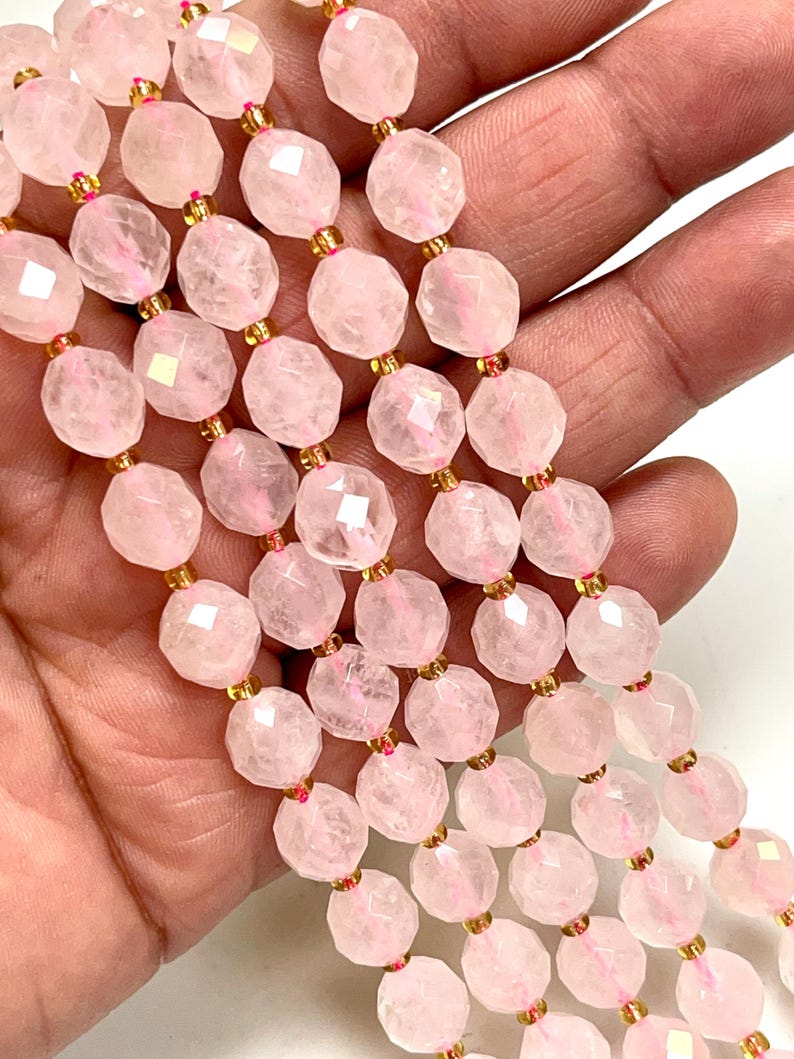 K&ouml;nnte beinhalten: Nahaufnahme von Str&auml;ngen facettierter, hellrosa Rosenquarzperlen. Die Perlen sind mit goldfarbenen Abstandshaltern aufgef&auml;delt. Die Perlen werden in einer Hand gehalten, was die zarte Farbe und Textur der Edelsteine zeigt.