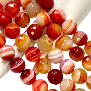 May include: A collection of round, polished gemstone beads in shades of red, orange, and cream. The beads have a banded pattern and a glossy finish, suggesting they are made of agate or a similar stone. The beads are likely for jewelry making.