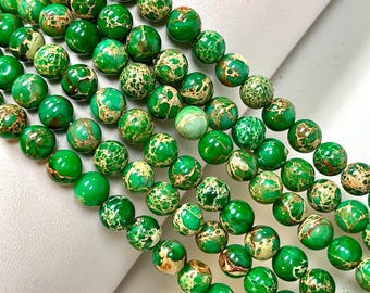 Puede incluir: Una hebra de cuentas de jaspe verde y dorado. Las cuentas son redondas y tienen un patrón moteado.