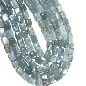 May include: Strands of aquamarine gemstone beads. The beads are a light blue colour with a square, slightly rounded shape. The beads are strung together, creating a flowing, organic pattern. The background is white.