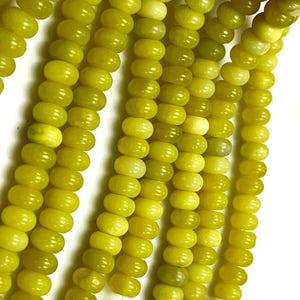May include: Close-up of strands of olive-green gemstone beads. The beads are oval-shaped and strung together, creating a textured surface. The beads have a glossy finish and vary slightly in color, ranging from light to dark olive green.