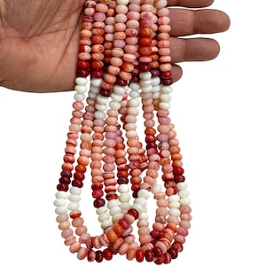 May include: Strands of colorful beads in shades of white, pink, and red. The beads are small and round, strung together to create a necklace or bracelet. The beads are held in a hand against a white background.