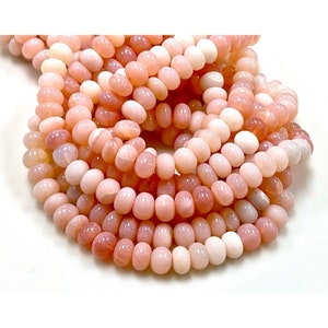 May include: A close-up of a strand of pink opal gemstone beads. The beads are oval-shaped and vary in shades of pink, from light blush to deeper rose. The beads are strung together, creating a textured and visually appealing display.