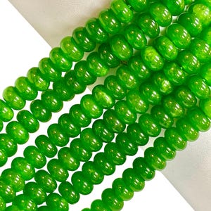 May include: Close-up of several strands of bright green beads. The beads are round with a slightly flattened shape and a glossy finish. The beads are strung together, creating a flowing, textured appearance. The background is white.