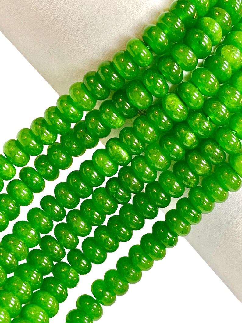 May include: Close-up of several strands of bright green beads. The beads are round with a slightly flattened shape and a glossy finish. The beads are strung together, creating a flowing, textured appearance. The background is white.