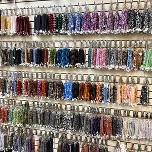Puede incluir: Una pared de exhibici&oacute;n de cuentas de piedras preciosas de varios colores, organizadas por color y enhebradas en alambre. Las cuentas se muestran en filas en una pared blanca con ganchos de metal.