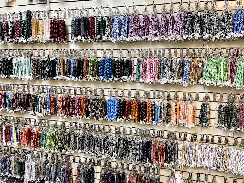 Puede incluir: Una pared de exhibici&oacute;n de cuentas de piedras preciosas de varios colores, organizadas por color y enhebradas en alambre. Las cuentas se muestran en filas en una pared blanca con ganchos de metal.