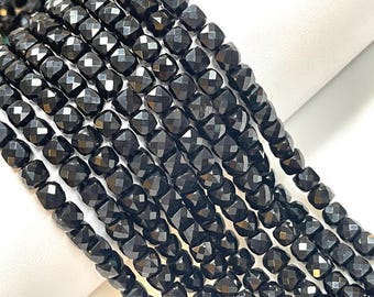 May include: Strands of black, faceted beads. Each bead is square with multiple facets, creating a sparkling effect. The beads are strung together, forming a continuous line. The background is white.