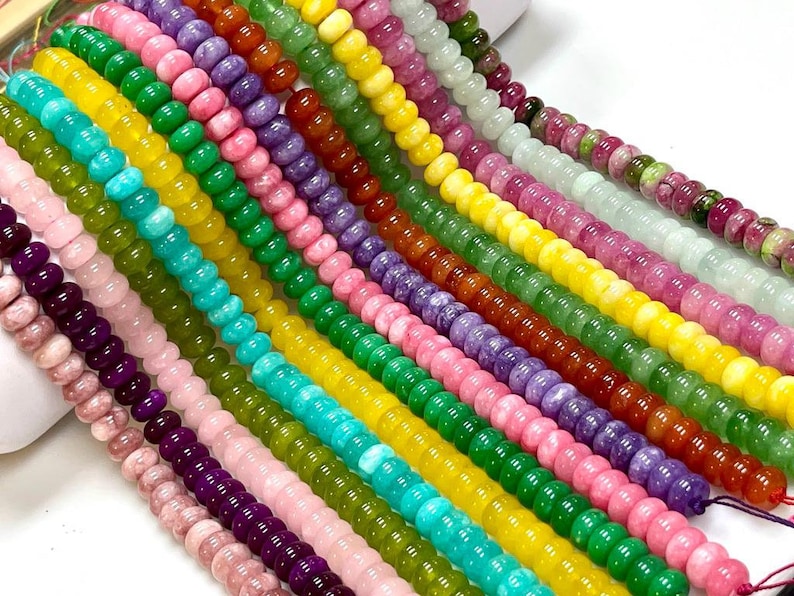 Op de afbeelding: Meerdere parallelle strengen van ronde, gladde en gepolijste edelsteenkralen. De kralen vertonen een levendig kleurenspectrum, waaronder roze, paars, donkerrood, felgroen, geel, turquoise en doorschijnend wit.