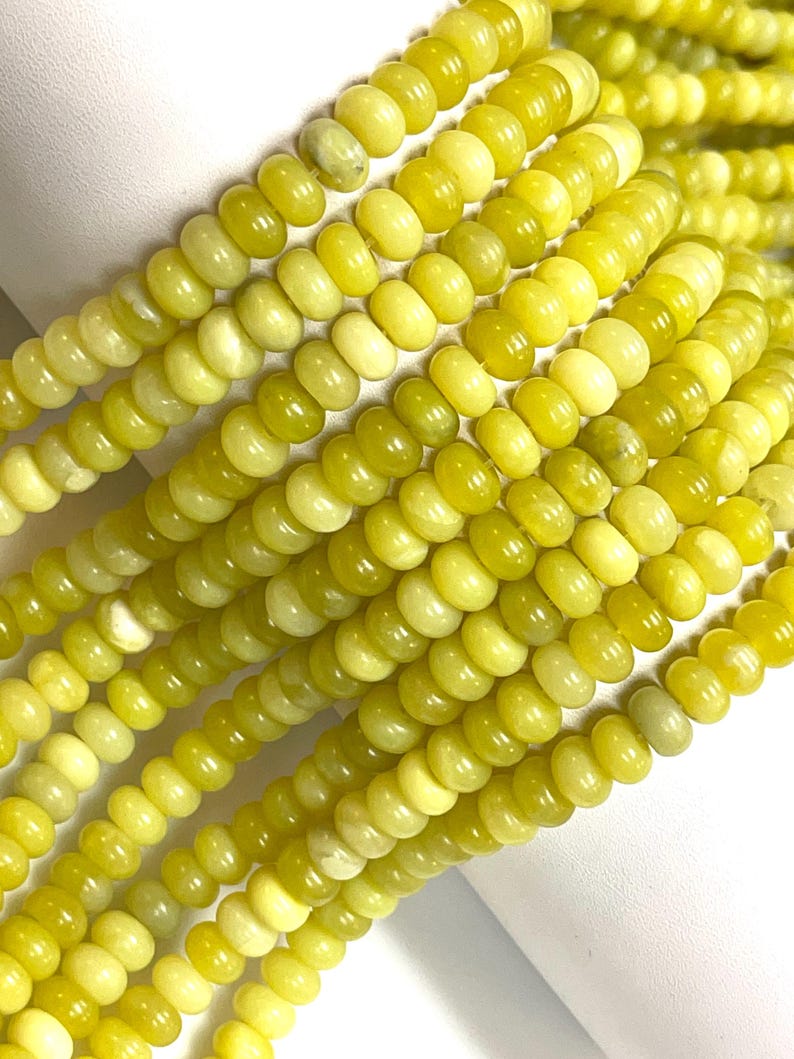 May include: Close-up of strands of yellow-green gemstone beads. The beads are a mix of light and darker shades of yellow-green, with a smooth, polished surface. The beads are strung together, creating a textured and colorful display.
