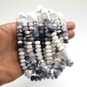 May include: Strands of white, gray, and black gemstone beads. The beads are disc-shaped and strung together, creating a layered effect. The beads are held in a hand against a white background. The beads have a speckled pattern.
