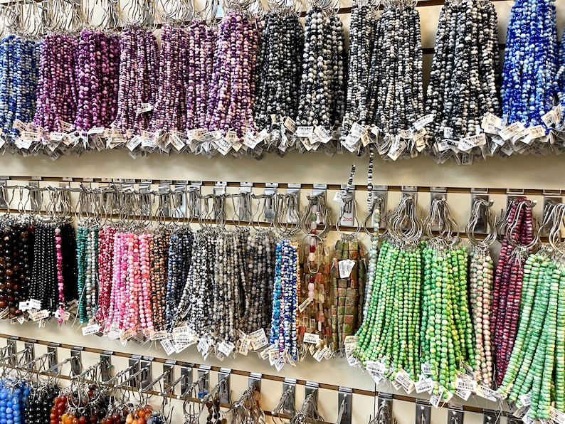Puede incluir: Una pared con una exposici&oacute;n de diferentes hileras de cuentas de colores, organizadas por color y colgadas de ganchos. Las cuentas est&aacute;n hechas de diferentes materiales, como vidrio, piedra y metal.