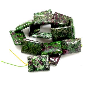 Puede incluir: Una colecci&oacute;n de cuentas rectangulares de rub&iacute; zoisita. Las cuentas son una mezcla de colores verde y burdeos, con un acabado pulido y brillante. Las cuentas est&aacute;n ensartadas con hilo amarillo y verde.