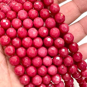 May include: Close-up of a strand of faceted, round beads in a vibrant red hue. The beads have a polished, glossy finish and are strung together, suggesting they are for jewelry making or crafting. The beads are held in a hand.