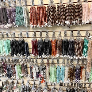 Puede incluir: Una pared de exposici&oacute;n con varias hebras de cuentas de diferentes colores y materiales, incluyendo madera, vidrio y piedra. Las cuentas est&aacute;n dispuestas en filas en ganchos, creando una exhibici&oacute;n colorida y visualmente atractiva.