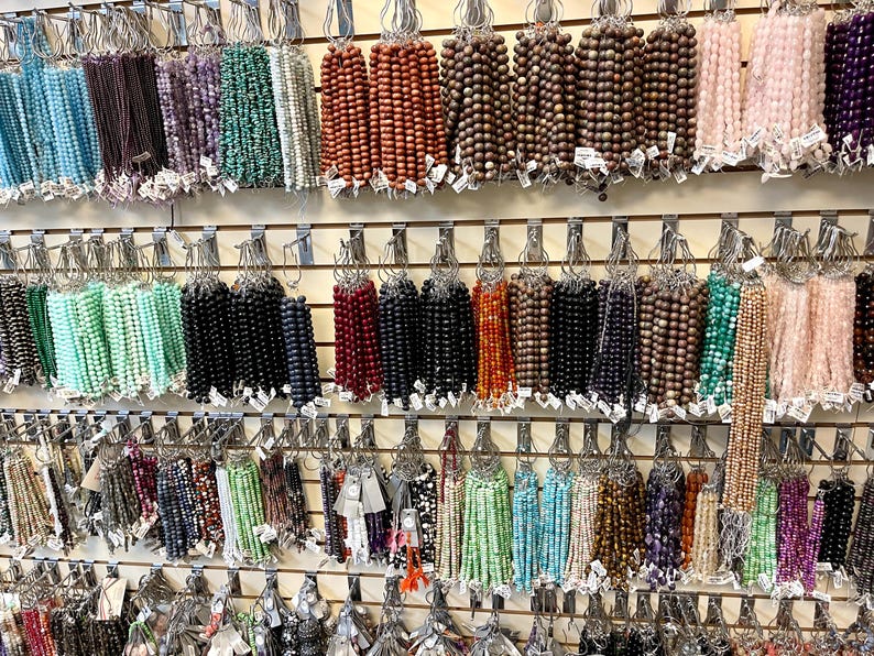 Puede incluir: Una pared de exposici&oacute;n con varias hebras de cuentas de diferentes colores y materiales, incluyendo madera, vidrio y piedra. Las cuentas est&aacute;n dispuestas en filas en ganchos, creando una exhibici&oacute;n colorida y visualmente atractiva.
