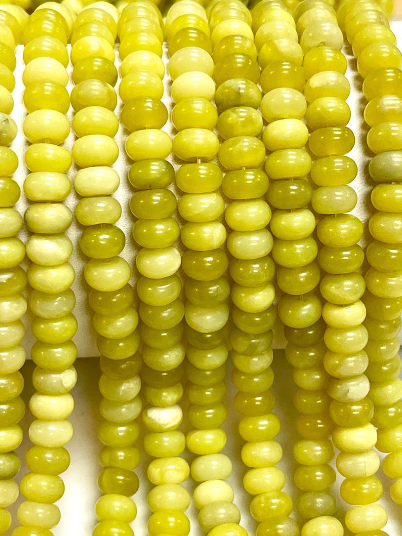 May include: Close-up of strands of yellow-green gemstone beads. The beads are round with a slightly flattened shape, and they have a smooth, polished surface. The color varies slightly, creating a subtle gradient effect. These beads are suitable for jewelry making.