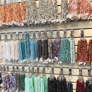 Puede incluir: Una pared de exhibici&oacute;n de cuentas de varios colores, organizadas por color y tipo. Las cuentas est&aacute;n enhebradas en cordones y colgadas de ganchos en una pared blanca. Las cuentas est&aacute;n hechas de diferentes materiales, incluyendo vidrio, piedra y madera.