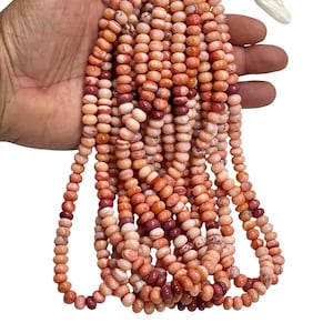 May include: Strands of coral-colored beads with varying shades of pink, peach, and burgundy. The beads are small, rounded, and strung together, creating a textured appearance. The beads are held in a hand against a white background.