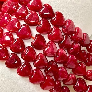 May include: A collection of polished, deep red heart-shaped beads arranged in multiple rows. Each bead features a smooth, glossy finish and a consistent heart silhouette, strung together to form a vibrant, uniform pattern of red gemstone-like shapes.