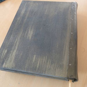 Puede incluir: Un diario de cuero con textura gris oscuro, encuadernado en cuero, con un marcador de cinta dorado.