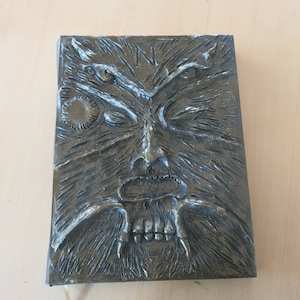 Puede incluir: Una cubierta de libro de color gris oscuro, texturizada, con una imagen esculpida en relieve de una criatura que parece un lobo que gru&ntilde;e, con una gran boca abierta y dientes afilados. Los ojos de la criatura est&aacute;n cerrados y la cubierta tiene un peque&ntilde;o dise&ntilde;o circular en la esquina superior izquierda.