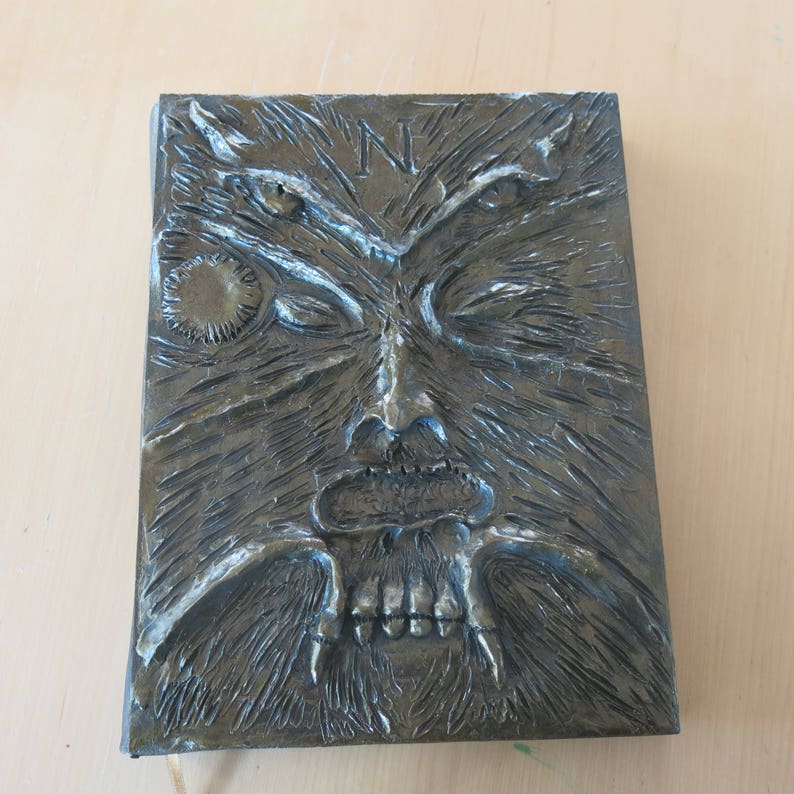 Puede incluir: Una cubierta de libro de color gris oscuro, texturizada, con una imagen esculpida en relieve de una criatura que parece un lobo que gru&ntilde;e, con una gran boca abierta y dientes afilados. Los ojos de la criatura est&aacute;n cerrados y la cubierta tiene un peque&ntilde;o dise&ntilde;o circular en la esquina superior izquierda.