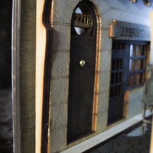Pode incluir: Um modelo de madeira de um edif&iacute;cio com uma porta etiquetada como "221B" e uma janela com o texto "LES SERVITEURS DE LA VILLE".