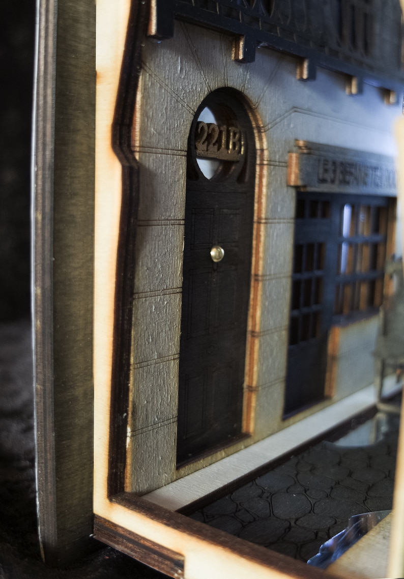 Pode incluir: Um modelo de madeira de um edif&iacute;cio com uma porta etiquetada como "221B" e uma janela com o texto "LES SERVITEURS DE LA VILLE".
