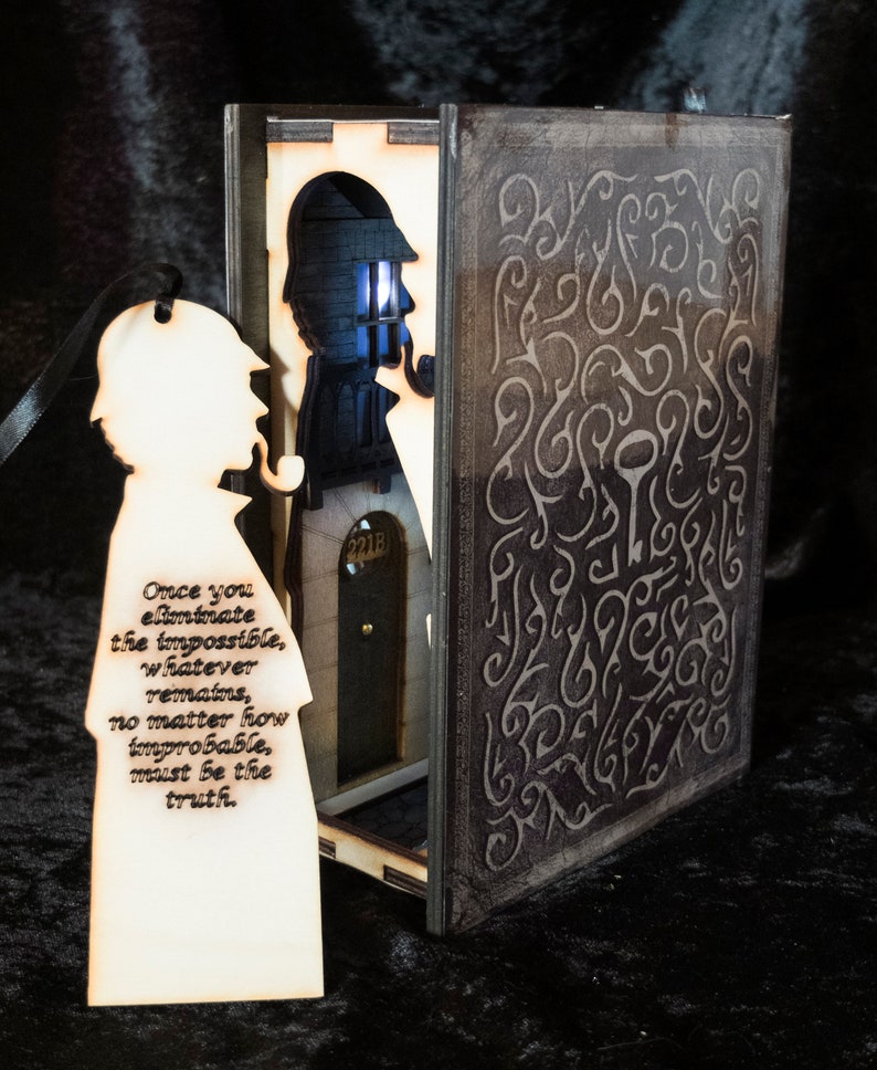 Pode incluir: Uma caixa de madeira em forma de livro com um recorte de Sherlock Holmes de p&eacute; em uma porta. A caixa est&aacute; aberta e o interior est&aacute; iluminado. A cita&ccedil;&atilde;o "Uma vez que voc&ecirc; elimina o imposs&iacute;vel, o que resta, n&atilde;o importa qu&atilde;o improv&aacute;vel seja, deve ser a verdade." est&aacute; gravada em um marcador de p&aacute;gina de madeira.