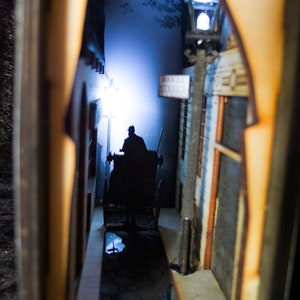 Pode incluir: Um modelo em miniatura de um beco escuro com a silhueta de uma pessoa em uma carruagem puxada por cavalos. O beco &eacute; iluminado por um poste de luz e a carruagem est&aacute; se movendo em dire&ccedil;&atilde;o &agrave; luz. O modelo &eacute; feito de madeira e tem uma apar&ecirc;ncia realista.